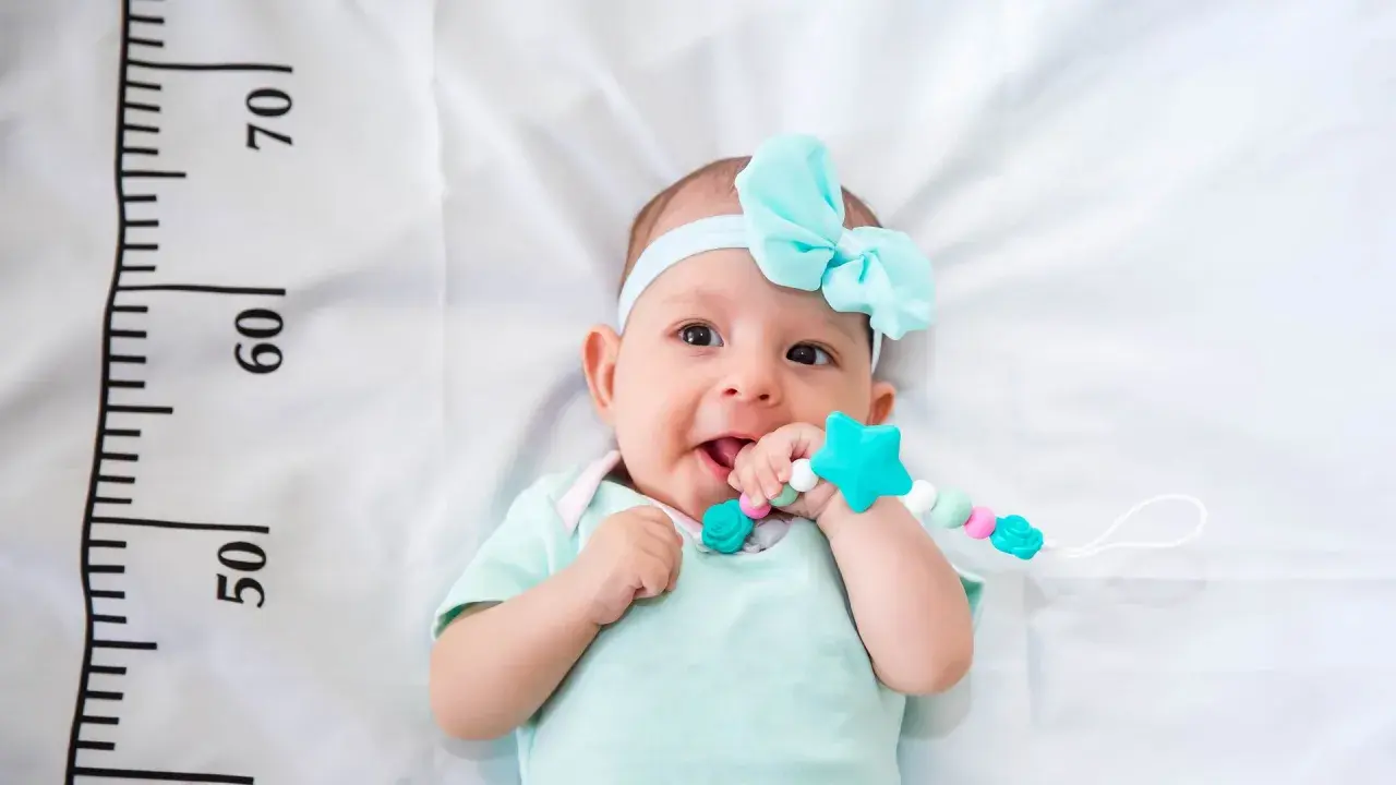 Un bebé sonriente con un chupete de estrella turquesa, con una cinta azul en la cabeza. A su lado, una regla marca el crecimiento, ¡un bebé perceptible!