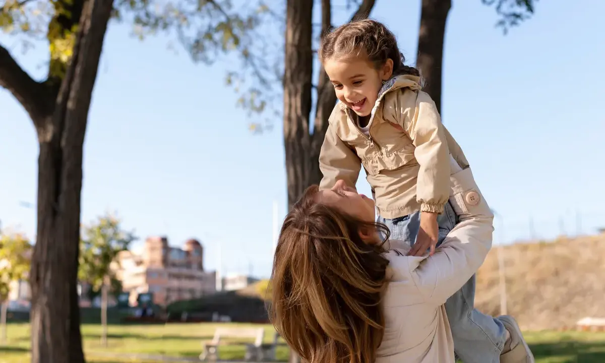 Madre levanta a su hija riendo, un momento lleno de palabras bonitas para niños y amor en un día soleado en el parque.