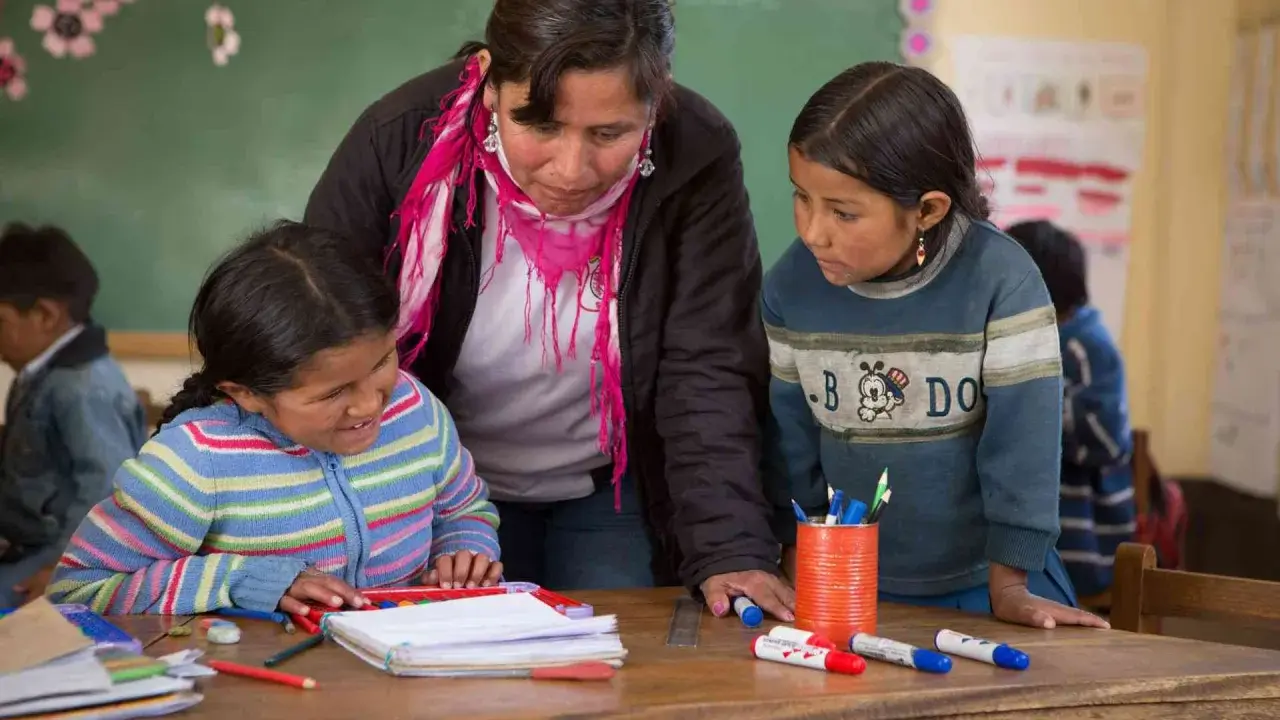 Maestra ayuda a dos niñas en un aula, promoviendo la importancia de las escuelas inclusivas.