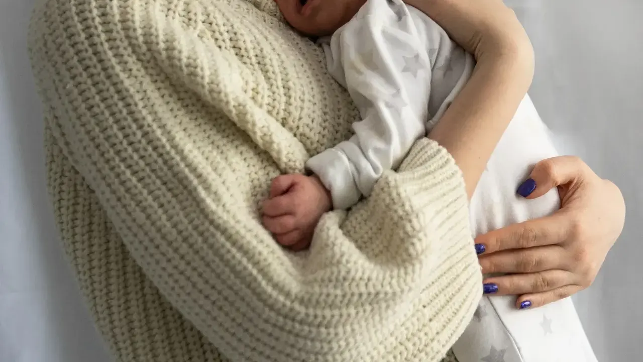 Bebé durmiendo acurrucado en brazos de su madre, envuelto en un suéter tejido. Un recordatorio de cuanto duerme un recién nacido.