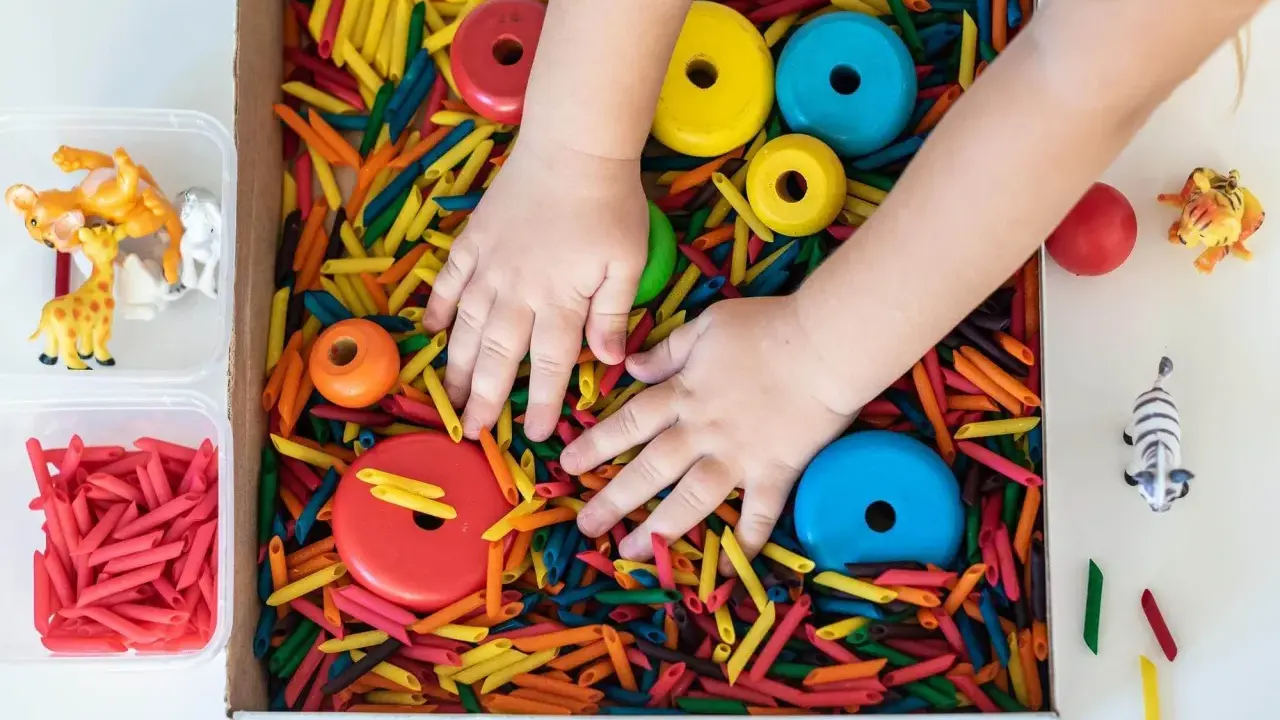 Manos de niño explorando un material sensorial de pasta de colores, con aros de madera y figuras de animales.