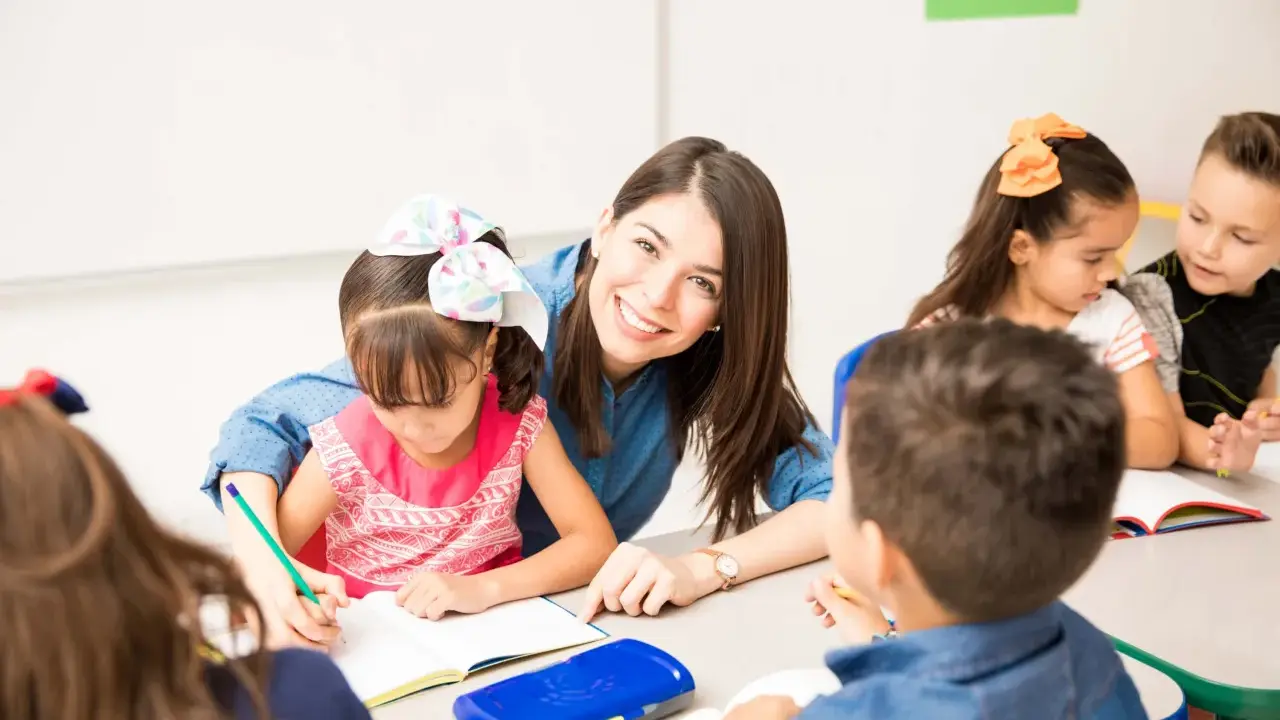 Maestra sonriente ayuda a una niña a escribir. Un aula llena de niños y frases bonitas para una profesora.