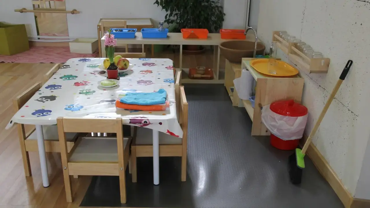 Mesa con mantel de búhos, toallas y frutas, lista para actividades en una casa de niños Montessori. Cerca, un lavabo y cubos para juegos.