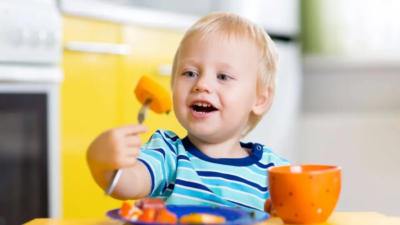 Niño rubio con tenedor y zanahoria, aprendiendo a comer solo. ¡Ayudame a hacerlo por mi mismo!