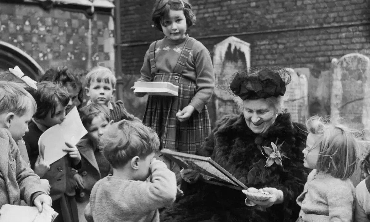 Maria Montessori joven, rodeada de niños, les muestra un libro. Una niña con tirantes sostiene una caja.