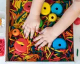 Manos de niño explorando un material sensorial de pasta de colores, con aros de madera y figuras de animales.