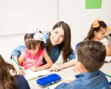 Maestra sonriente ayuda a una niña a escribir. Un aula llena de niños y frases bonitas para una profesora.