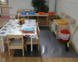 Mesa con mantel de búhos, toallas y frutas, lista para actividades en una casa de niños Montessori. Cerca, un lavabo y cubos para juegos.