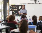 Maestro sonriente dirige una clase, los alumnos usan tabletas para su evaluación en casa.