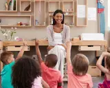 Niños en un aula **Waldorf Montessori** levantan las manos para responder a su maestra sonriente.