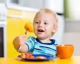 Niño rubio con tenedor y zanahoria, aprendiendo a comer solo. ¡Ayudame a hacerlo por mi mismo!