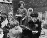 Maria Montessori joven, rodeada de niños, les muestra un libro. Una niña con tirantes sostiene una caja.