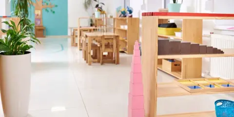Un aula Montessori 0-3 años con mesas bajas, estanterías de madera y materiales didácticos como la torre rosa.