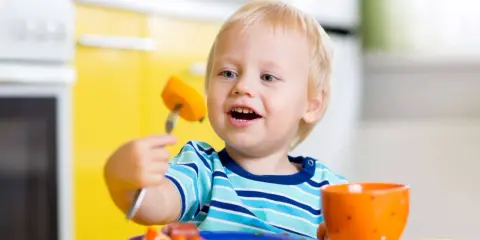 Niño rubio con tenedor y zanahoria, aprendiendo a comer solo. ¡Ayudame a hacerlo por mi mismo!