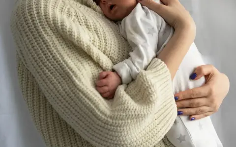 Bebé durmiendo acurrucado en brazos de su madre, envuelto en un suéter tejido. Un recordatorio de cuanto duerme un recién nacido.