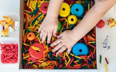 Manos de niño explorando un material sensorial de pasta de colores, con aros de madera y figuras de animales.