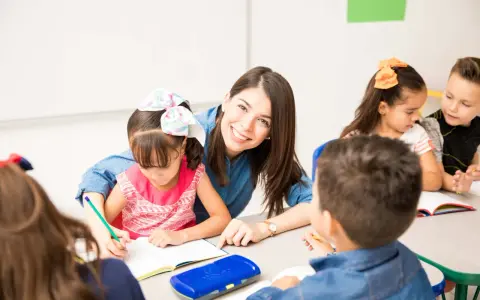 Maestra sonriente ayuda a una niña a escribir. Un aula llena de niños y frases bonitas para una profesora.