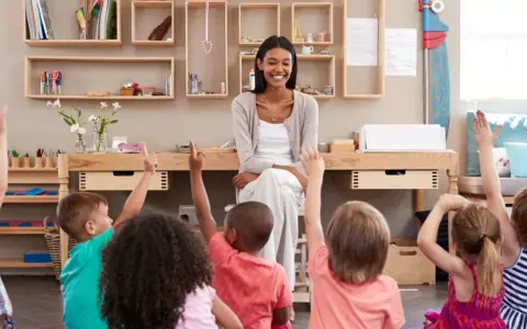 Niños en un aula **Waldorf Montessori** levantan las manos para responder a su maestra sonriente.