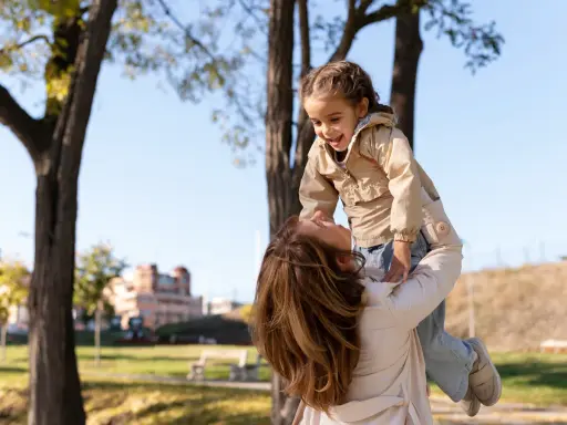 Madre levanta a su hija riendo, un momento lleno de palabras bonitas para niños y amor en un día soleado en el parque.