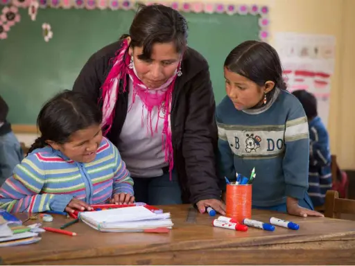 Maestra ayuda a dos niñas en un aula, promoviendo la importancia de las escuelas inclusivas.