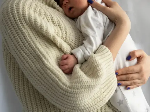 Bebé durmiendo acurrucado en brazos de su madre, envuelto en un suéter tejido. Un recordatorio de cuanto duerme un recién nacido.