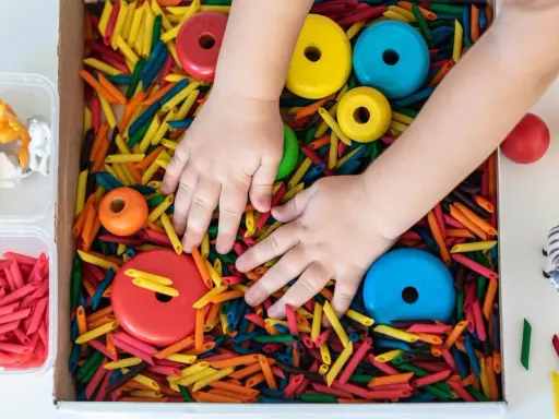 Manos de niño explorando un material sensorial de pasta de colores, con aros de madera y figuras de animales.