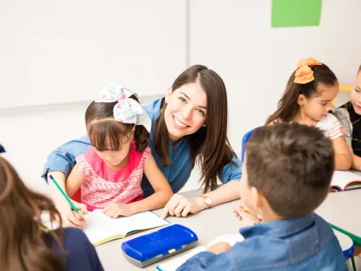 Maestra sonriente ayuda a una niña a escribir. Un aula llena de niños y frases bonitas para una profesora.
