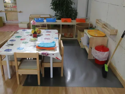 Mesa con mantel de búhos, toallas y frutas, lista para actividades en una casa de niños Montessori. Cerca, un lavabo y cubos para juegos.