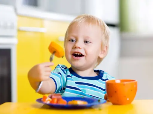 Niño rubio con tenedor y zanahoria, aprendiendo a comer solo. ¡Ayudame a hacerlo por mi mismo!