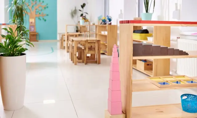 Un aula Montessori 0-3 años con mesas bajas, estanterías de madera y materiales didácticos como la torre rosa.