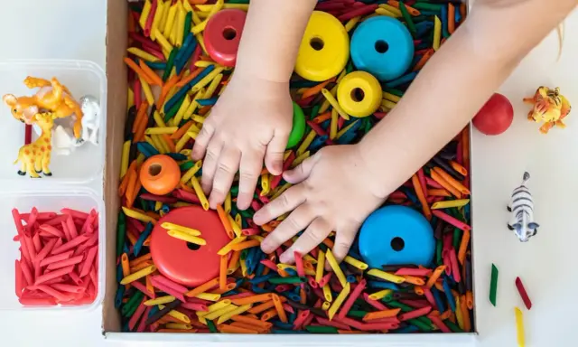 Manos de niño explorando un material sensorial de pasta de colores, con aros de madera y figuras de animales.