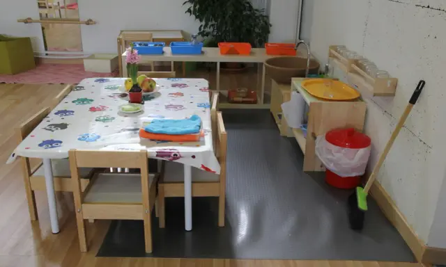Mesa con mantel de búhos, toallas y frutas, lista para actividades en una casa de niños Montessori. Cerca, un lavabo y cubos para juegos.