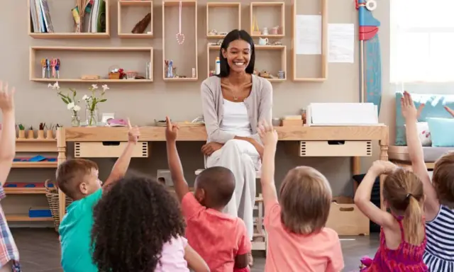 Niños en un aula **Waldorf Montessori** levantan las manos para responder a su maestra sonriente.