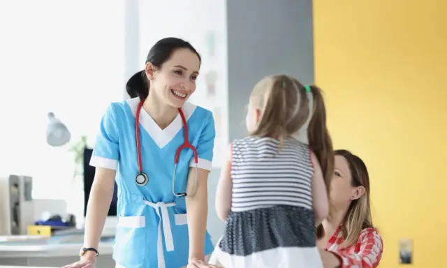 Doctora sonríe a niña y madre, ofreciendo atención educativa en un entorno médico.