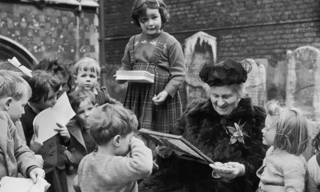 Maria Montessori joven, rodeada de niños, les muestra un libro. Una niña con tirantes sostiene una caja.