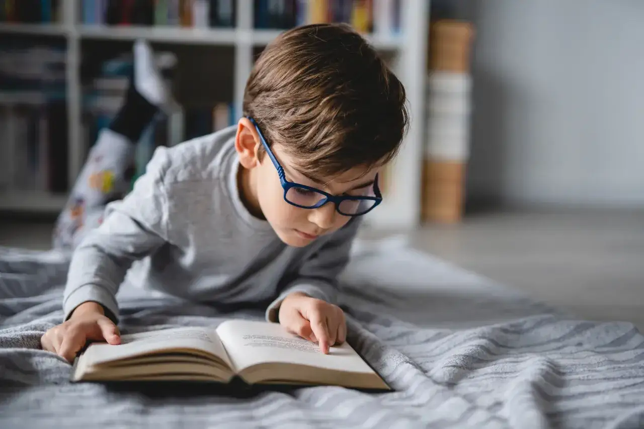 Ni&ntilde;o con gafas lee un libro abierto, rodeado de estanter&iacute;as llenas de libros por edades.