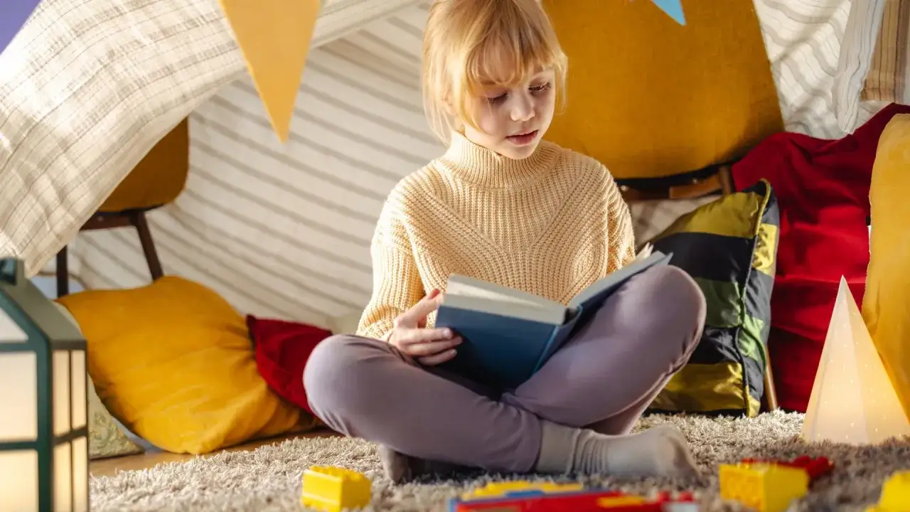 Ni&ntilde;a rubia lee un libro de misterio para ni&ntilde;os de 10 a 12 a&ntilde;os, acurrucada en un fuerte de mantas.