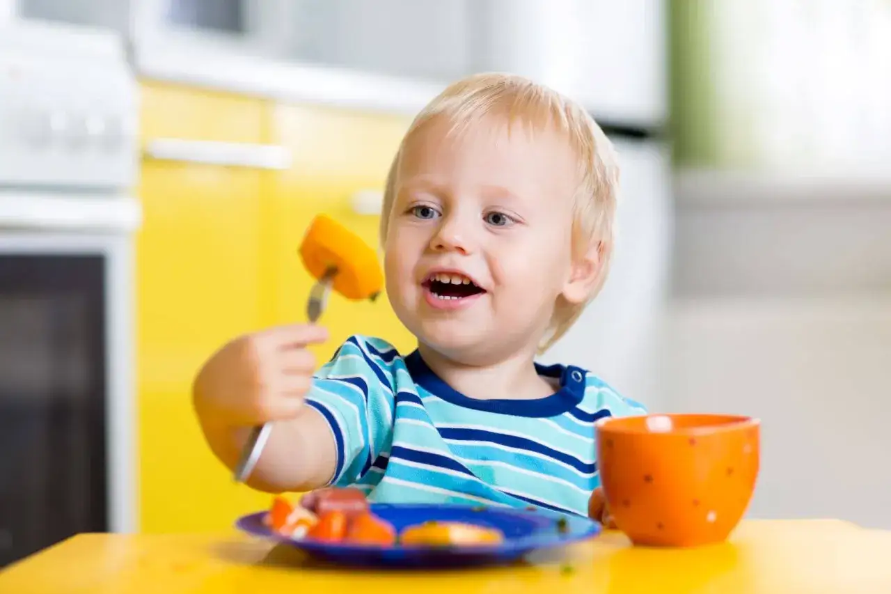 Niño rubio de 0-3 años en un aula Montessori, comiendo zanahoria con tenedor.