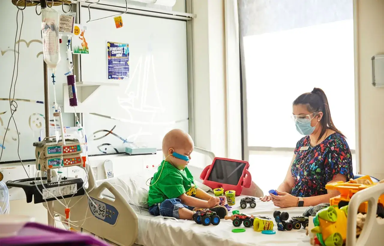Ni&ntilde;o en cama de hospital, rodeado de juguetes y equipo m&eacute;dico, mientras una enfermera observa. El aprendizaje perceptivo se fomenta a trav&eacute;s del juego y la interacci&oacute;n.