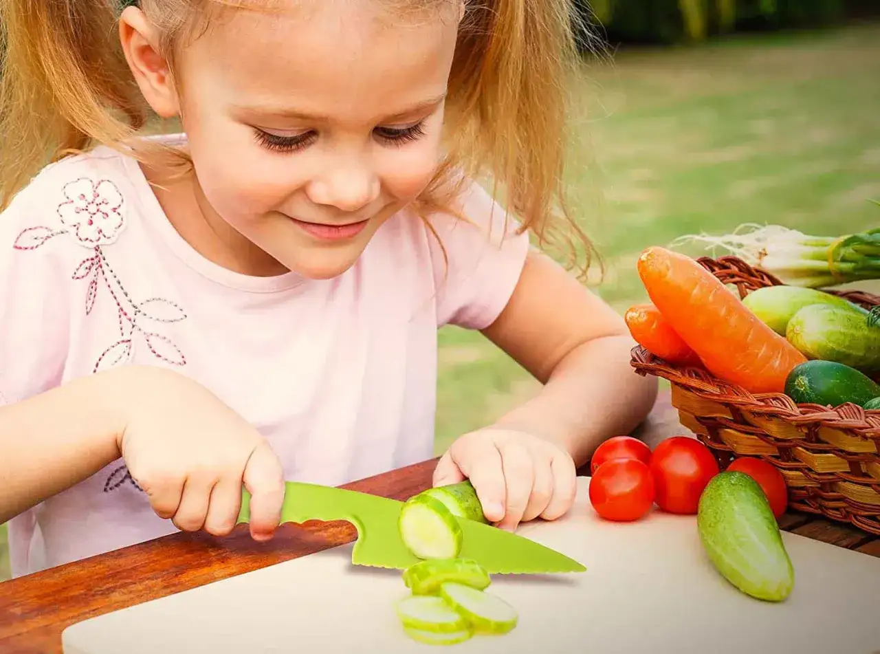 Ni&ntilde;a sonriente practicando vida pr&aacute;ctica Montessori, cortando pepino con cuchillo de pl&aacute;stico. Verduras frescas en cesta.