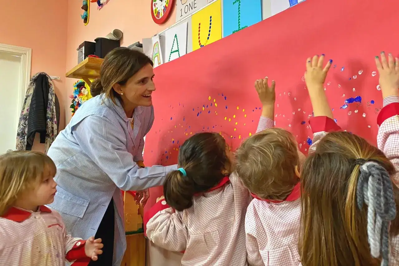 Maestra sonríe mientras niños pintan en pared. Inspiración para frases para profesores de infantil.