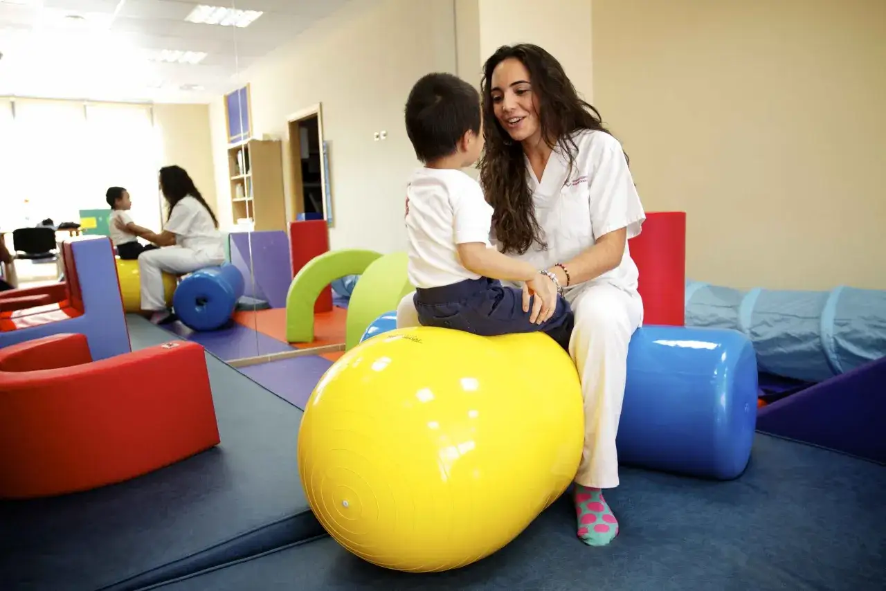 Terapeuta interact&uacute;a con un ni&ntilde;o en una sala de terapia, fomentando el desarrollo del cerebro infantil con materiales l&uacute;dicos.