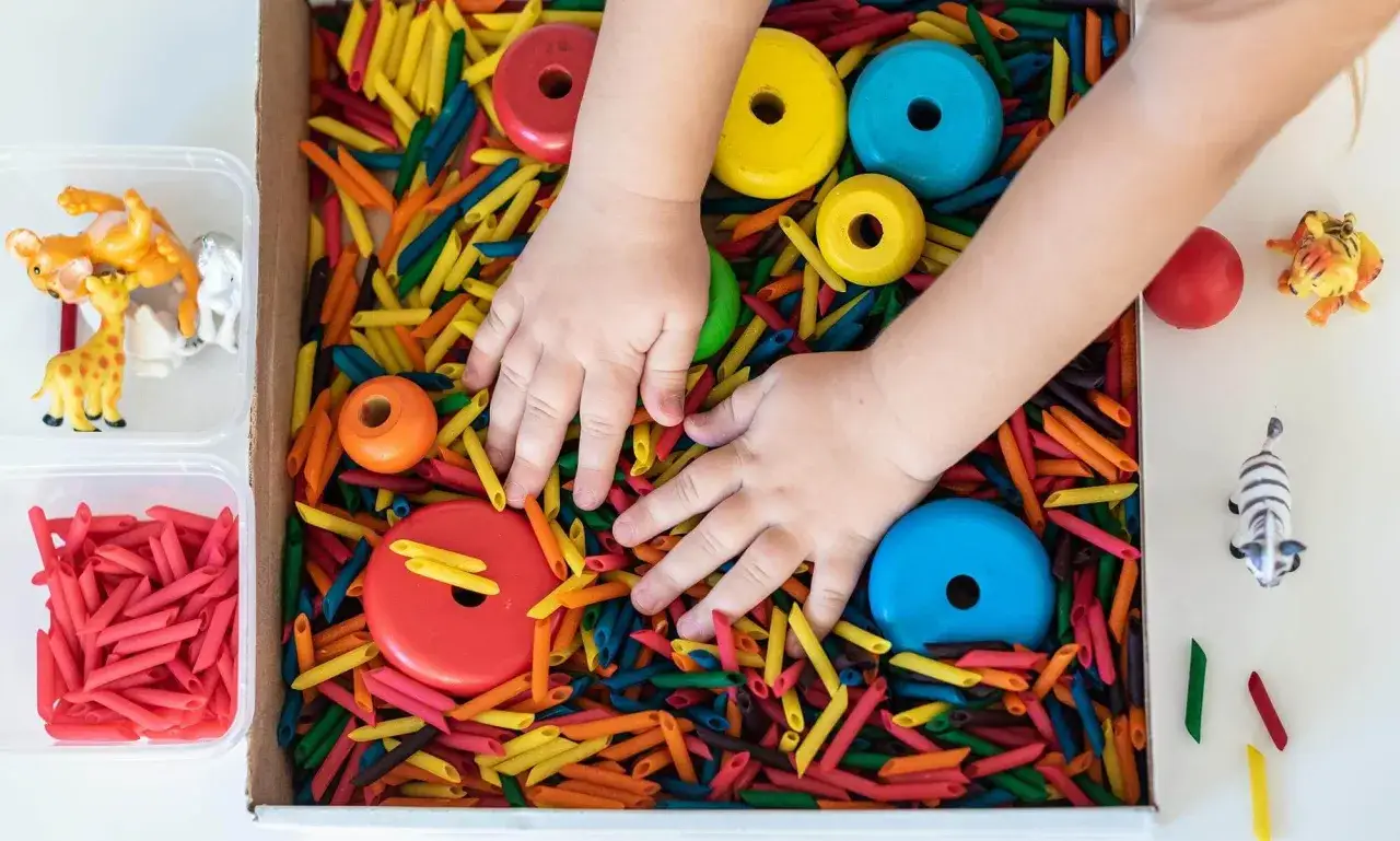 Manos de ni&ntilde;o explorando un material sensorial de pasta de colores con aros de madera y figuras de animales.