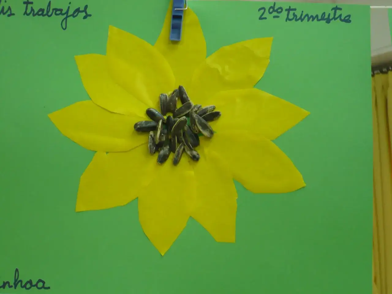 Portada primavera infantil: un girasol de papel amarillo con semillas en el centro, colgado con una pinza azul.