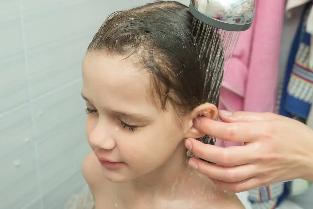 Ni&ntilde;a se lava el pelo bajo la ducha, mientras una mano limpia la suciedad detr&aacute;s de las orejas.