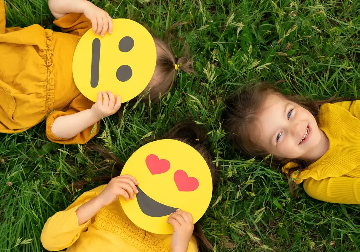 Ni&ntilde;as jugando con caras de emojis para imprimir, expresando alegr&iacute;a y amor en la hierba.