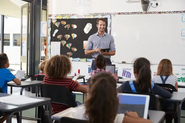 Maestro sonriente dirige una clase, los alumnos usan tabletas para su evaluación en casa.